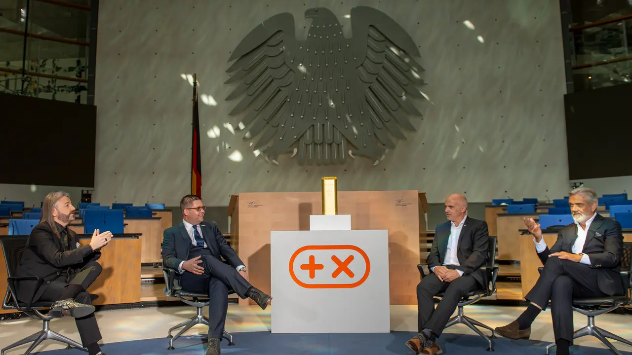 Vier Gesprächspartner in der TV-Sendung „Marken im Fokus“, in der Mitte die Trophäe, dahinter der Bundesadler