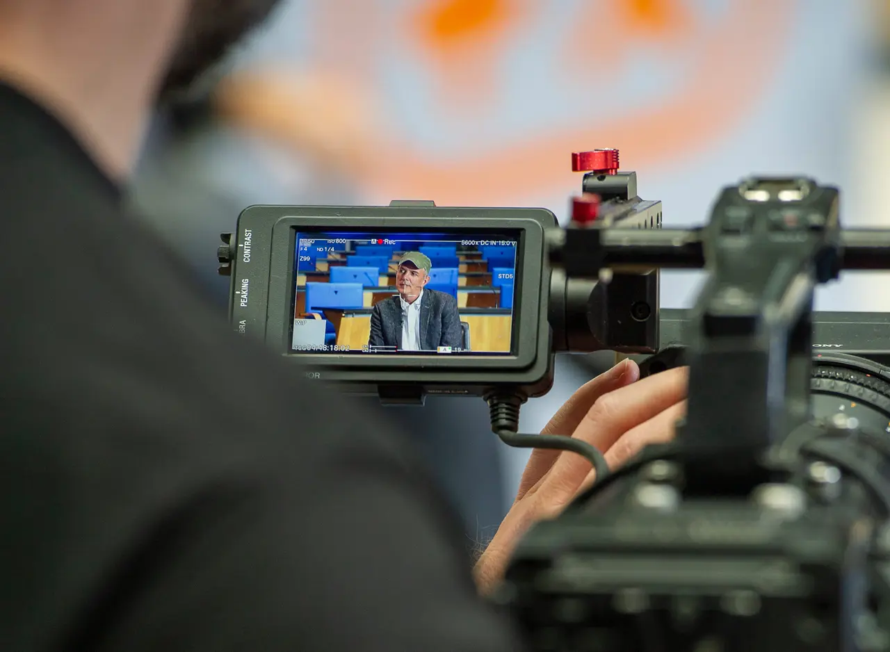 Kameramonitor zeigt einen Interviewgast vor den blauen Sitzreihen des ehemaligen Plenarsaals