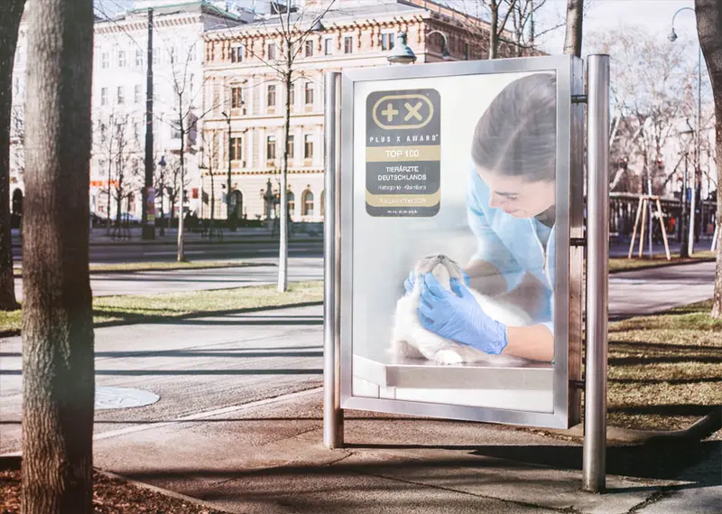 Freistehender Schaukasten am Strassenrand bei Tageslicht; das Plakat zeigt eine Tierärztin bei der Untersuchung einer Katze und das Plus X Award Siegel „Top 100 Tierärzte Deutschlands“