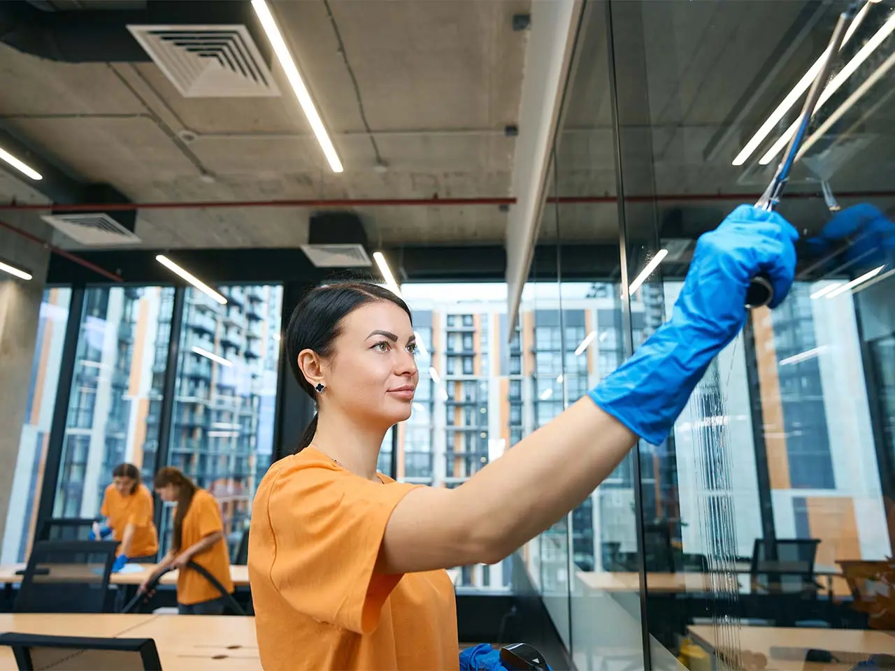 Reinigungskräfte in orangefarbenen Shirts säubern ein Großraumbüro, vorn wird eine Glaswand abgezogen